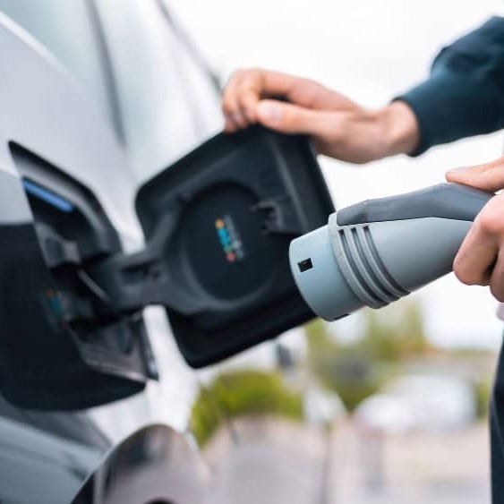 Instalación de cargadores de coches eléctricos 1 Primer plano de un usuario sujetando un conector de carga robusto (negro y gris) a punto de insertarlo en un coche eléctrico negro. Ilustra la facilidad de uso y la calidad del hardware de un wallbox instalado por Ojoalvatio.